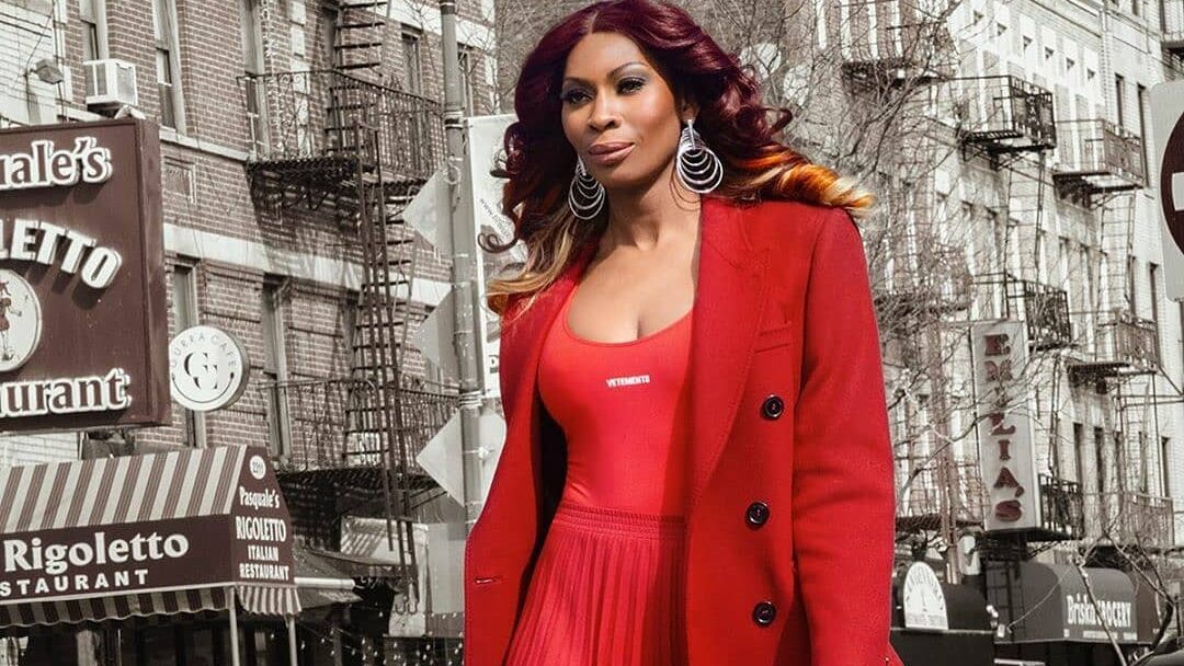 Dominique Jackson walks confidently in NYC, wearing a custom all-red outfit with a high slit and matching coat.