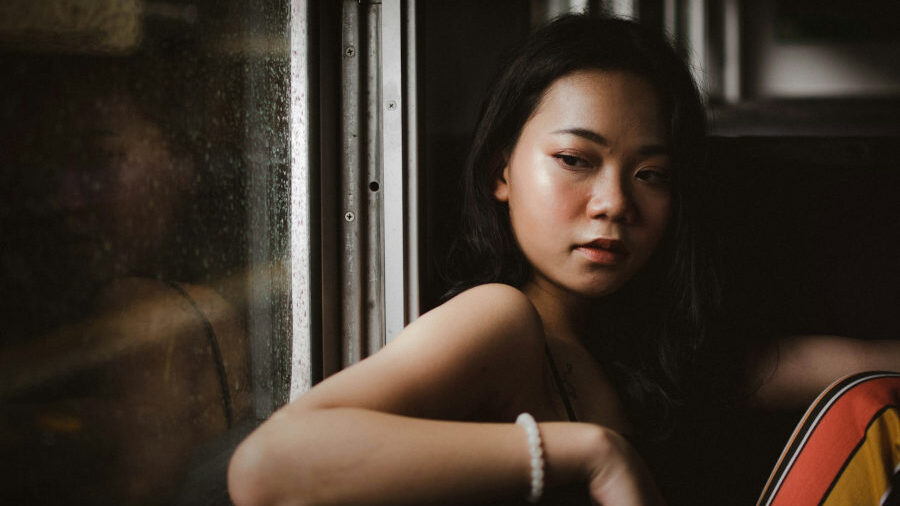 Woman with long black hair gazes out a rain-covered window, leaning quietly on her arm in soft natural light.