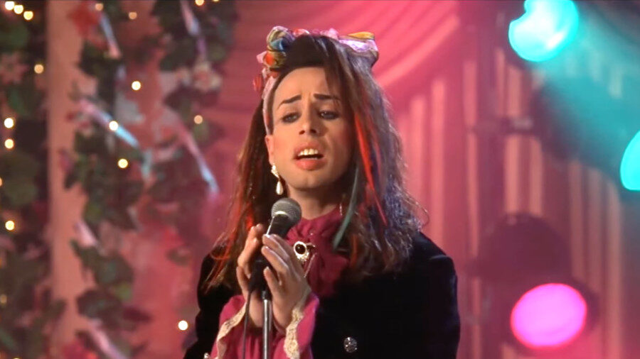 Alexis Arquette on stage in a colorful bow and dark outfit, singing into a mic.