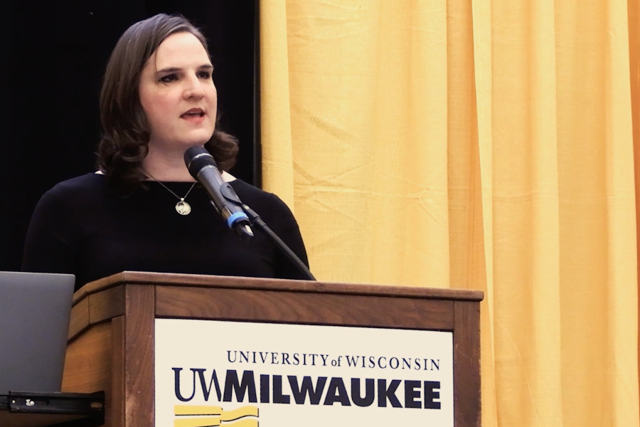 Ashley Altadonna speaks at a podium during UWM’s Lavender Graduation 2023, with a curtain and UWM logo in background.