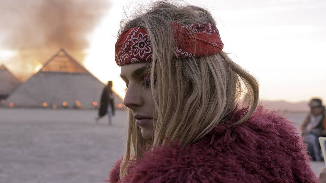 Andreja Pejić stands in the Burning Man desert in 2016, wearing a red bandana and faux fur coat, fire glowing behind her.
