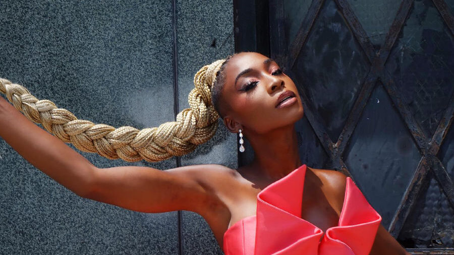 Angelica Ross poses in a sculptural pink dress with a long braided ponytail and drop earrings, leaning against a dark wall.