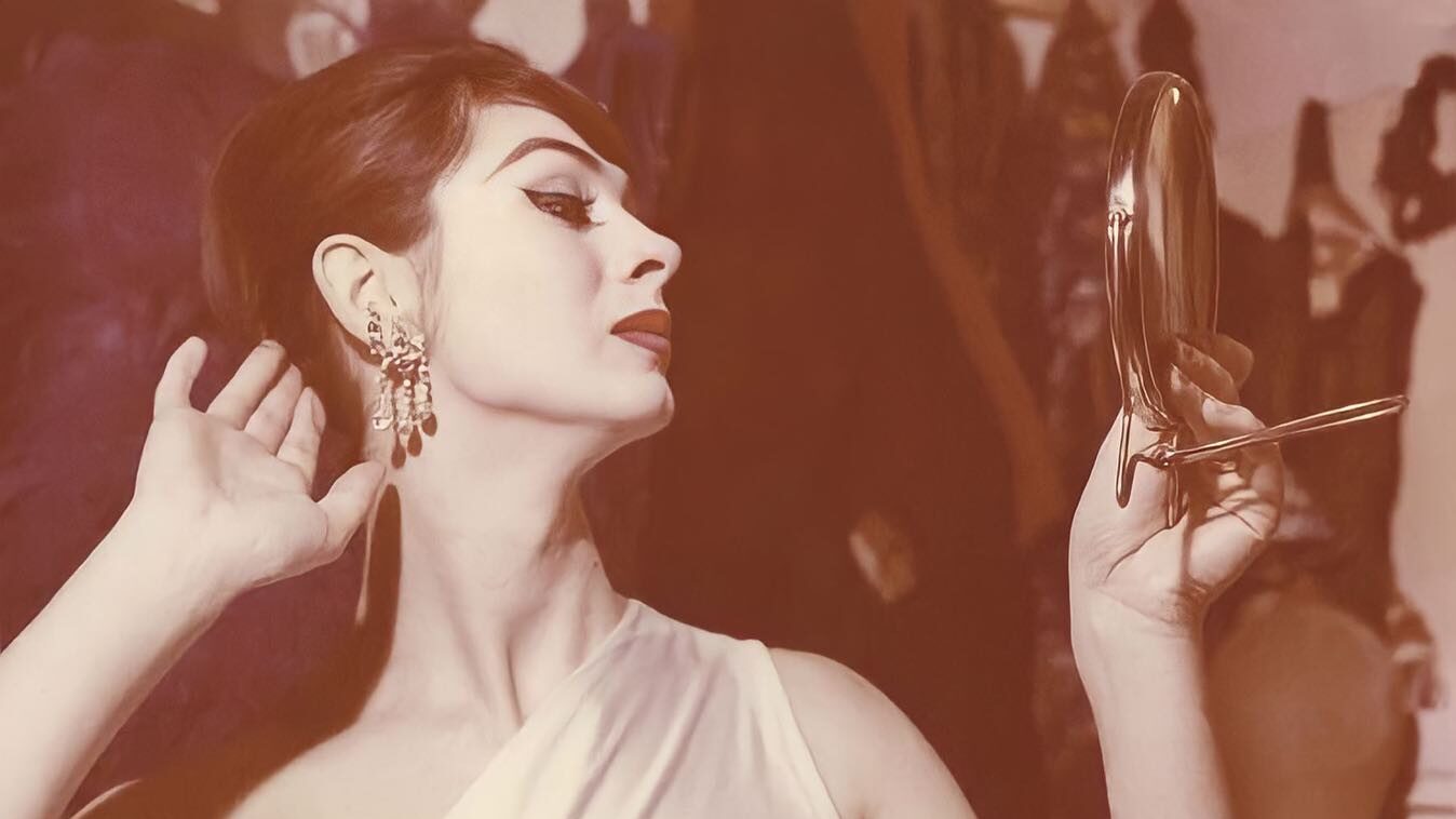 Vintage photo of April Ashley admiring her reflection, wearing statement earrings and a white gown.