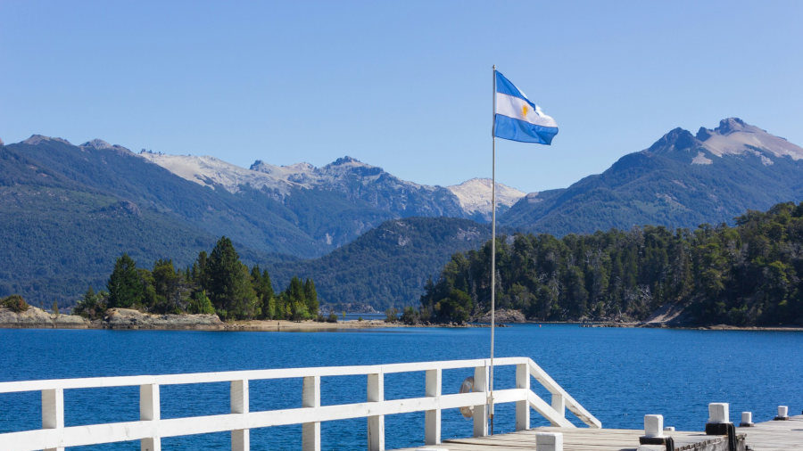 Argentine flag on a pier overlooking a blue lake and forested mountains in Patagonia under clear blue sky.