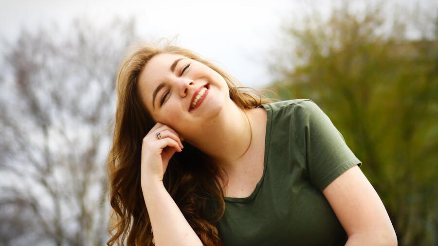 Woman smiling with eyes closed outdoors, leaning her head on her hand with trees blurred in the background.