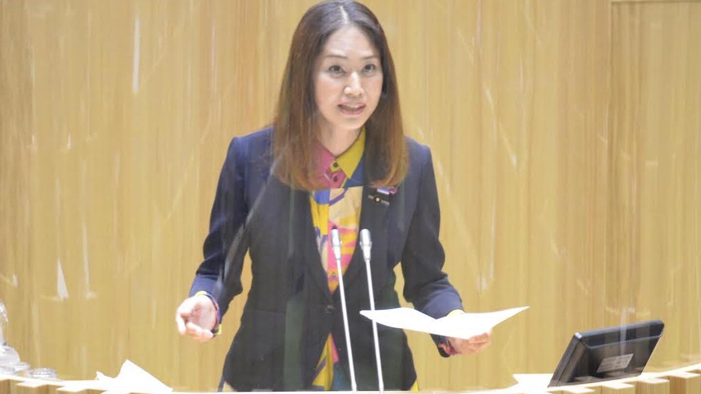 Ayako Fuchigami mid-speech, wearing a dark jacket and colorful shirt at the assembly