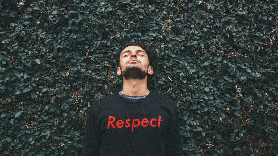 Person in a black sweater with “Respect” in red letters, standing in front of a dense green wall of ivy with eyes closed.