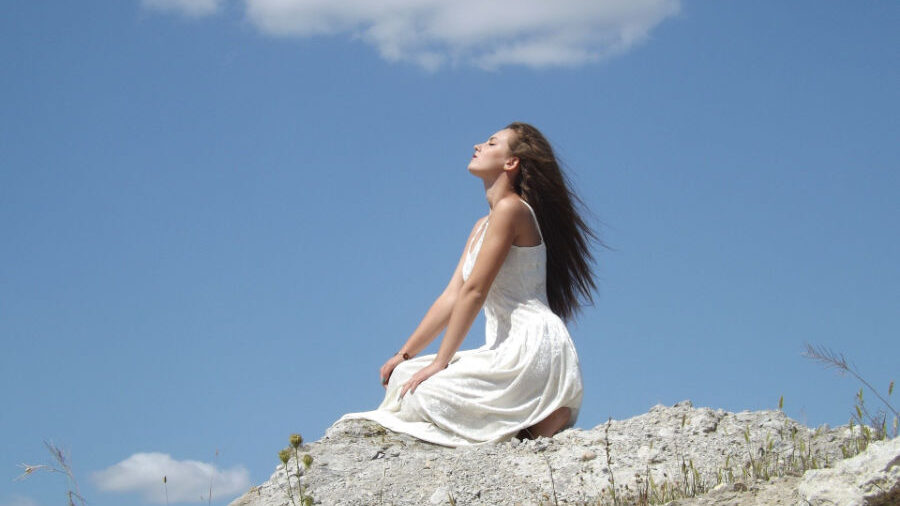 Woman in a flowing white dress, seated on rocky terrain, breathing in the fresh air.