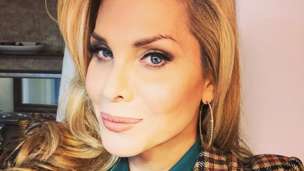 Candis Cayne souriante en selfie, boucle d’oreille, veste à carreaux et chemise verte, cheveux ondulés.