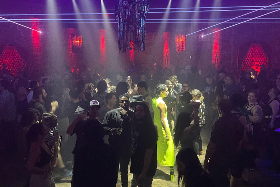 A large crowd dances under purple laser lights at The Castle gay bar, with disco balls and a party vibe.