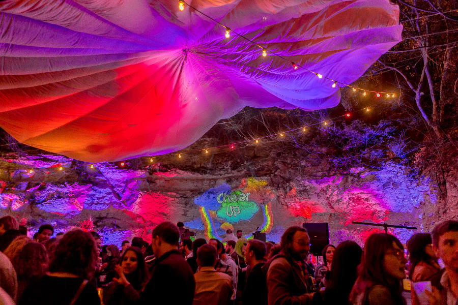 Colorful night scene at Cheer Up Charlie’s with a crowd under fabric canopy and string lights, rainbow projection on rocky wall.