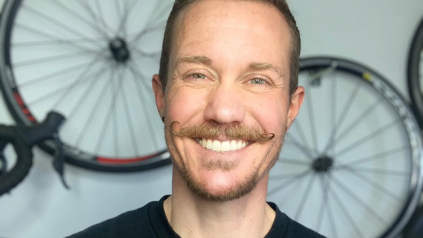 Chris Mosier souriant devant des vélos, portant un t-shirt noir “Activist Athlete” chez lui.