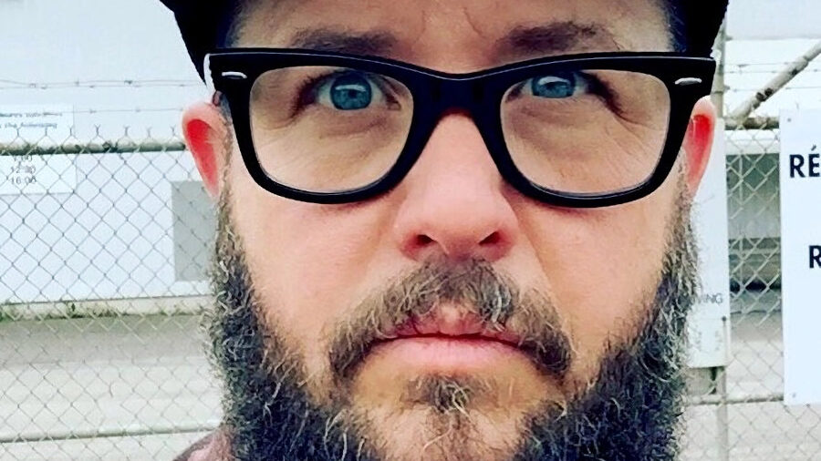 Cooper Lee Bombardier avec barbe, lunettes et casquette, portant une chemise à carreaux en extérieur.