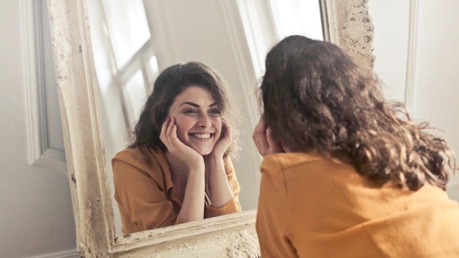 Happy woman with curly hair gazing lovingly at her reflection, embracing her identity and inner confidence.