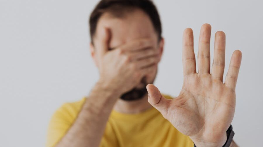 A man in a yellow shirt covers his eyes with one hand and extends the other toward the camera in a stop gesture.