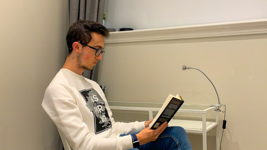 Young man wearing glasses and a white sweatshirt reads a book in a minimalist room with a small desk and lamp.