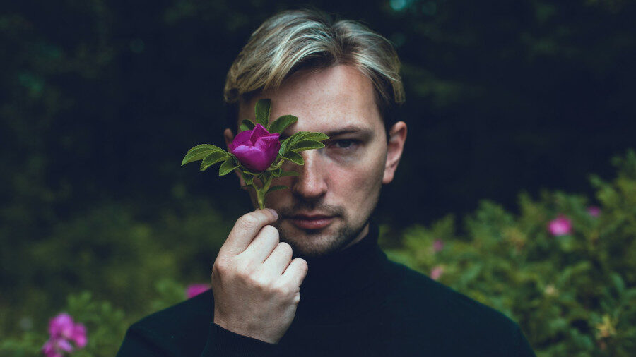 A blond-haired person in a black turtleneck holds a vibrant pink flower over one eye, with greenery in the background.