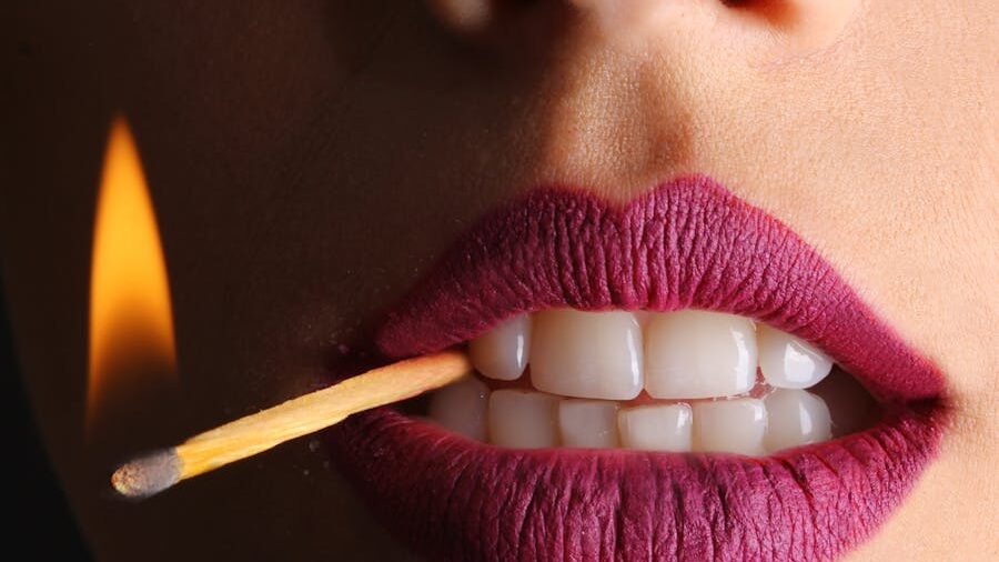 Artistic photo of purple lips biting a burning match, with visible flame and high-contrast studio lighting.