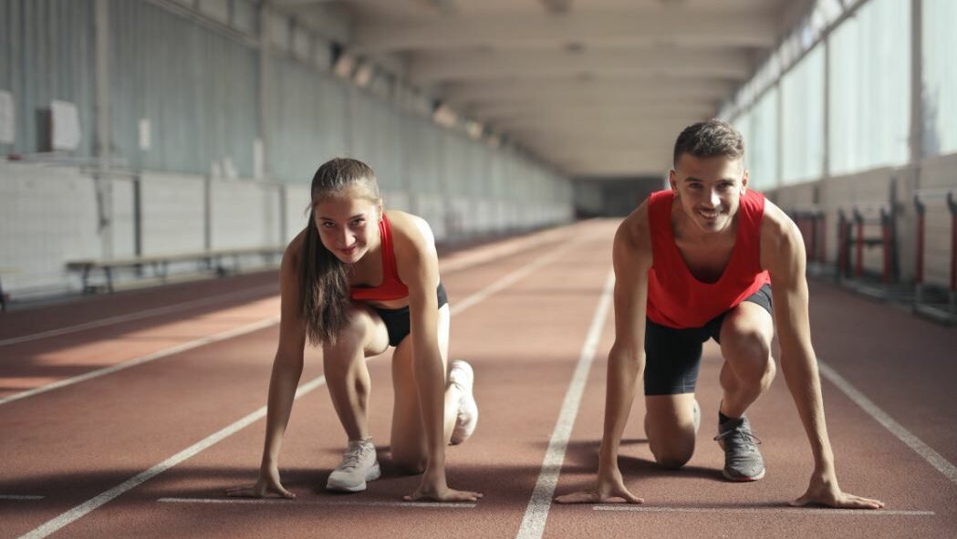 Two athletes in red jerseys crouched at a starting position on an indoor track.