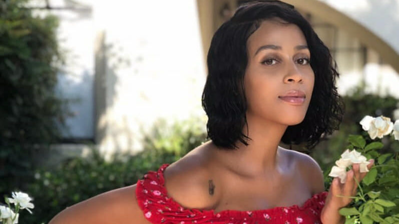 Isis King posing among white flowers in a garden, wearing a red crop top and jeans.