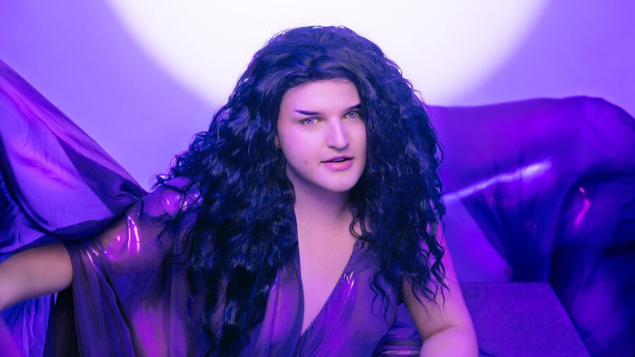 Jae XO in a sheer purple outfit with long curly hair, seated under violet light.