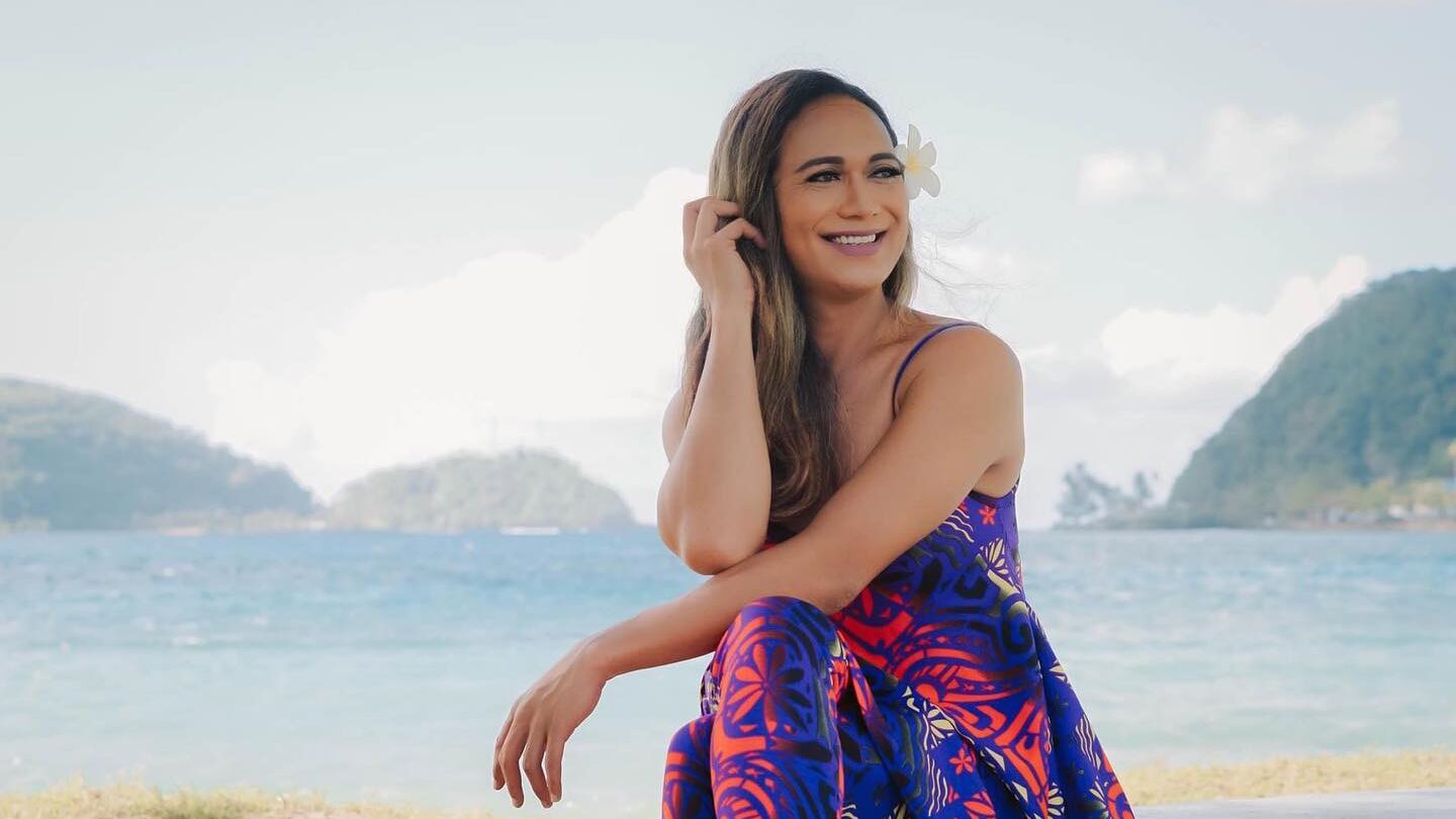 Jaiyah Saelua assise sur l’herbe près de l’océan aux Samoa, portant une robe colorée.