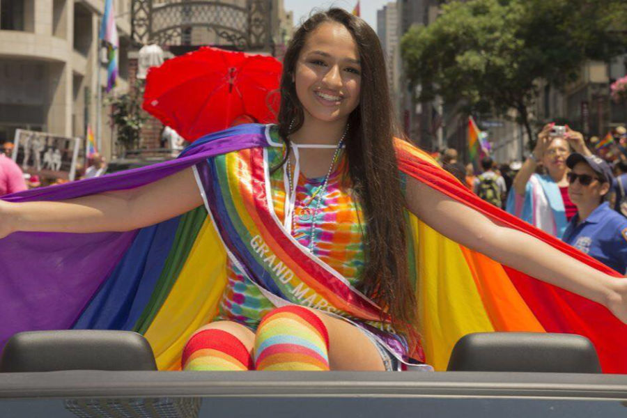Jazz Jennings, wearing a rainbow dress and cape, sits on a car as Grand Marshal at a Pride parade, smiling with arms outstretched.