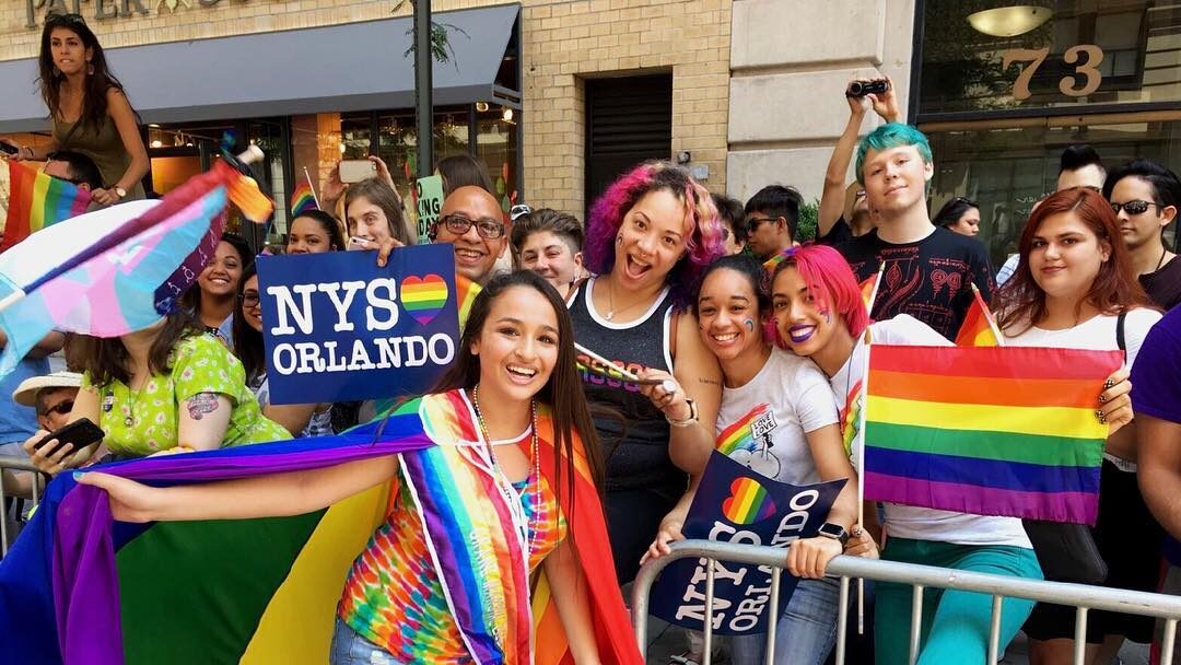 Jazz Jennings smiling in rainbow attire as Grand Marshal at NYC Pride 2016, surrounded by joyful participants holding Pride flags and signs.