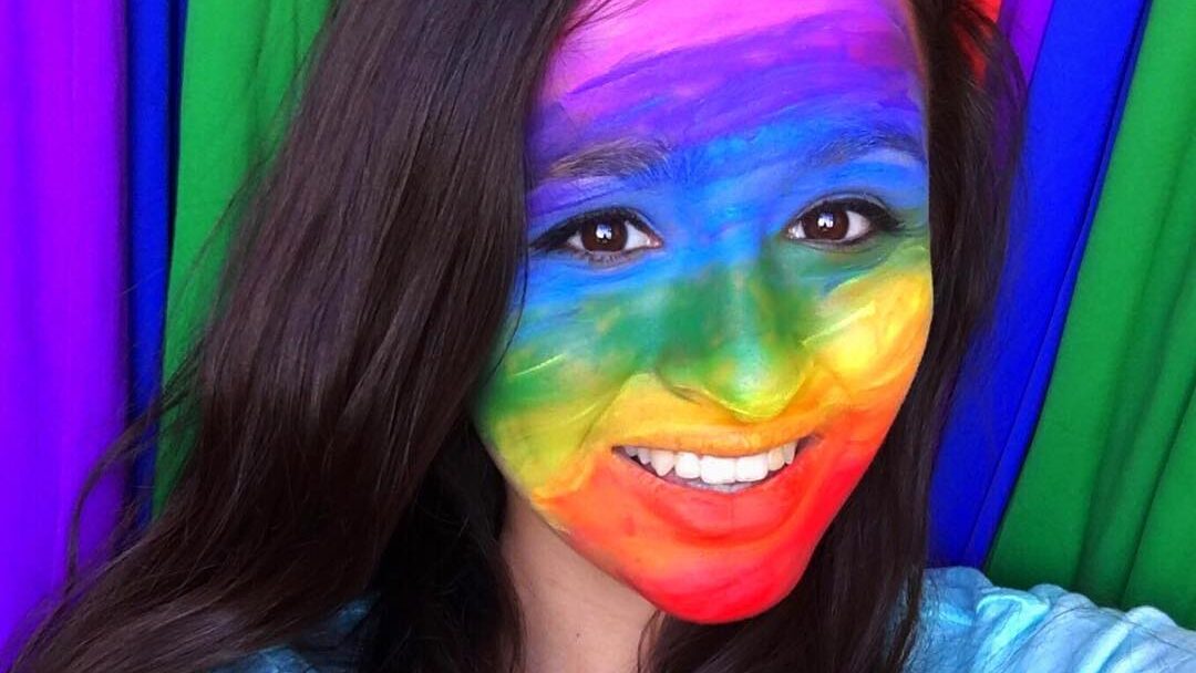 Jazz Jennings smiling in front of a rainbow backdrop, her face painted in rainbow colors for Pride.