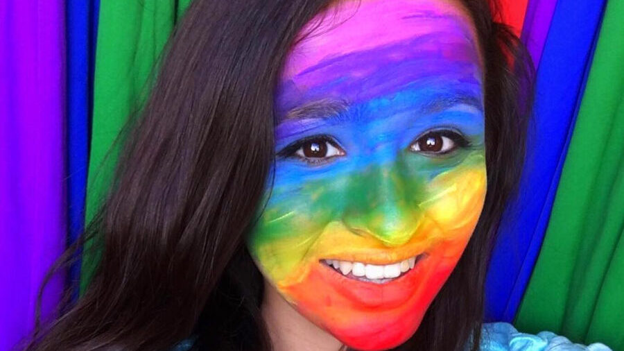 Jazz Jennings smiling in front of a rainbow backdrop, her face painted in rainbow colors for Pride.