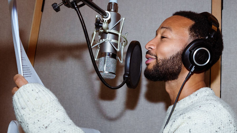 John Legend wearing headphones and reading from a script into a studio microphone.