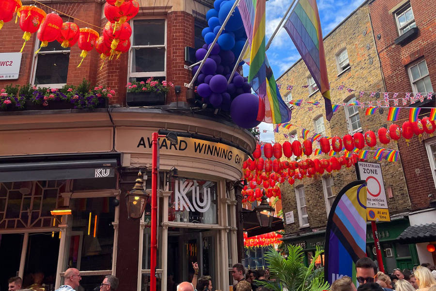 KU Bar façade with rainbow flags, red lanterns and balloons during a crowded London Pride celebration.