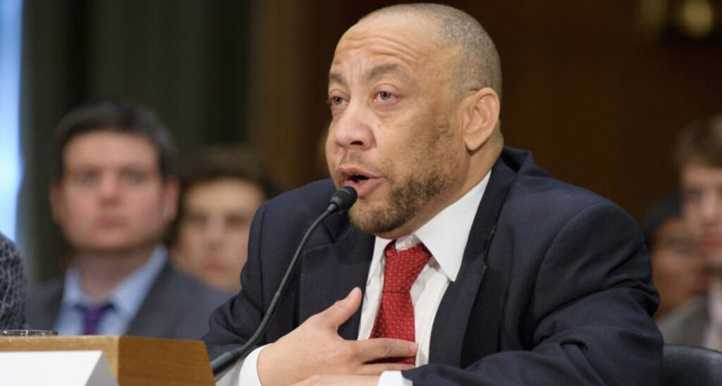 Kylar Broadus speaks into a microphone during a U.S. Senate hearing, seated at a wooden desk with others in the background.