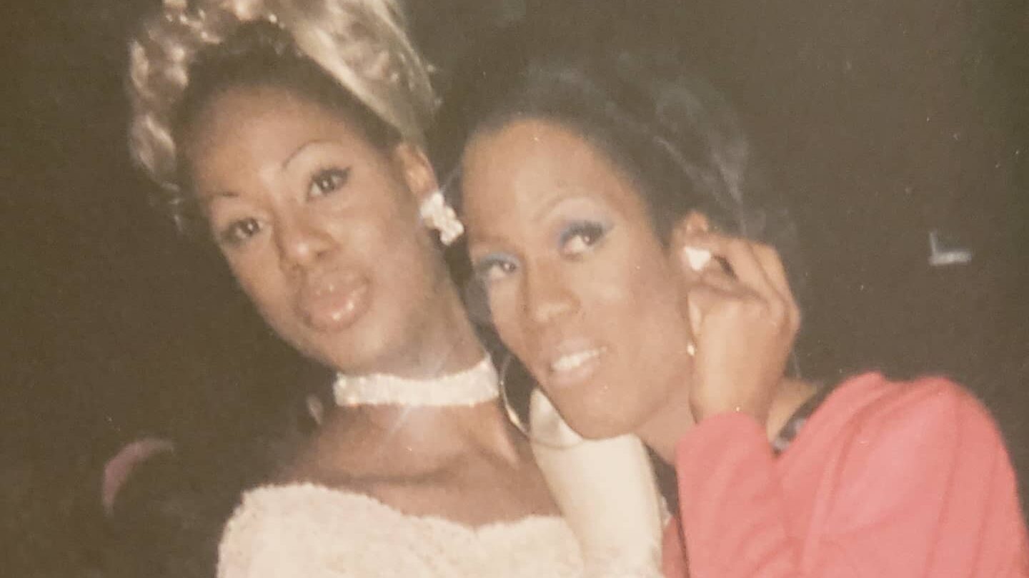 Une photo vintage montre Laverne Cox et Shequida Hall déguisées pour une soirée d’Halloween en 1999. Laverne porte une robe blanche à épaules dénudées et une perruque blonde, tandis que Shequida est habillée en rose avec un maquillage glamour. Elles posent côte à côte, souriantes, sur une photo aux tons chauds légèrement fanés, prise au club Roxy à New York.