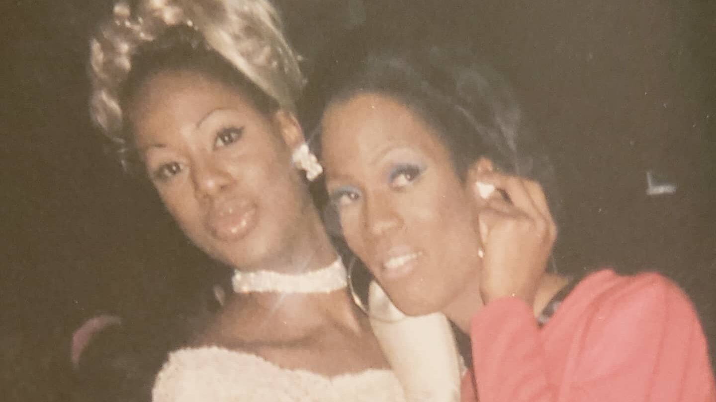 A vintage photograph of Laverne Cox and Shequida Hall dressed up for a Halloween party in 1999. Laverne wears a white off-shoulder dress and blonde wig, while Shequida sports a pink outfit and glamorous makeup. They pose closely, smiling in the warm-toned, slightly faded photo taken at the Roxy nightclub in NYC.
