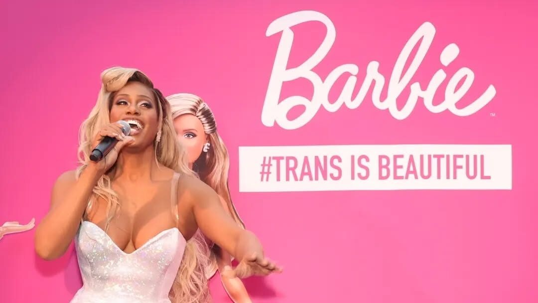 Laverne Cox sourit dans une combinaison blanche scintillante, un micro à la main, devant un fond rose Barbie avec sa poupée posée à côté d’elle. Le slogan “#TRANS IS BEAUTIFUL” s’affiche derrière elle, célébrant cette collaboration révolutionnaire.