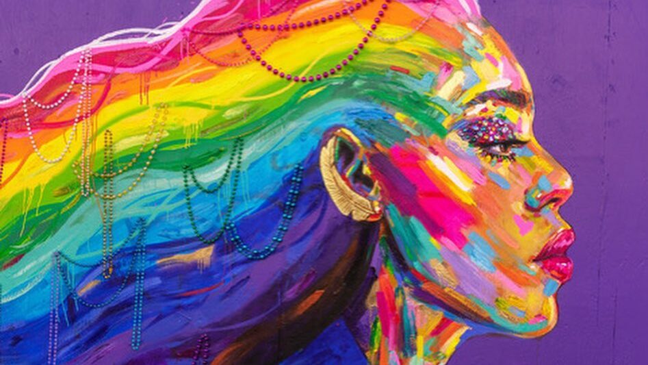 A vibrant mural of Laverne Cox painted in rainbow colors on a purple background, with her long hair flowing in rainbow waves adorned with pearls. The portrait was created by artist Kate Tova in honor of Spirit Day, symbolizing strength, pride, and solidarity with LGBTQ+ youth.