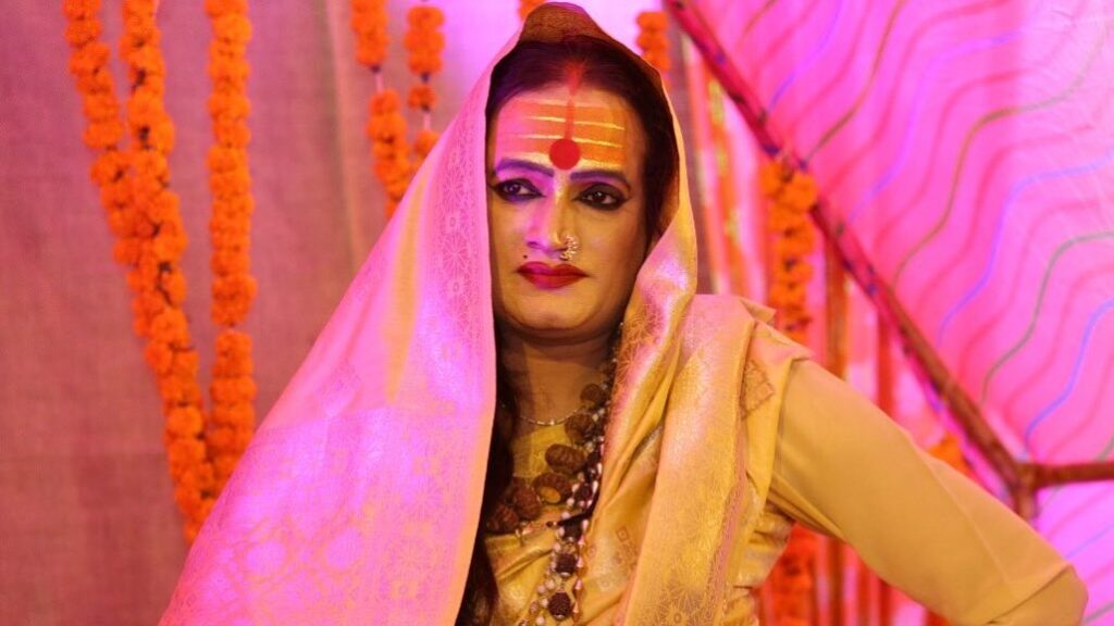 Laxmi Narayan Tripathi seated cross-legged in a yellow and gold sari, under pink lights.