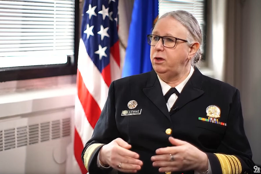 Admiral Rachel Levine speaking during an interview, wearing her uniform with medals, seated beside U.S. and federal flags.