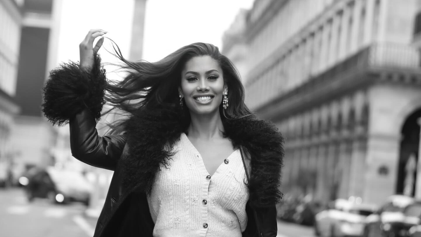 Leyna Bloom marche avec assurance au milieu d’une rue parisienne, portant un manteau bordé de fourrure sur un ensemble en maille, un sac à main à la main, avec l’architecture haussmannienne en arrière-plan.