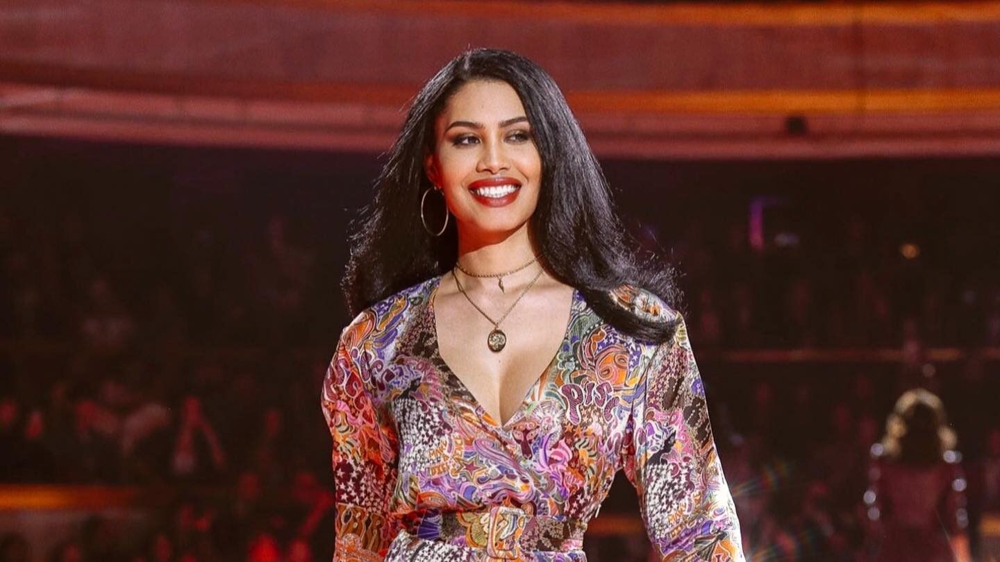 Leyna Bloom défile sur un podium illuminé, vêtue d’une robe colorée à motifs, souriante face au public.