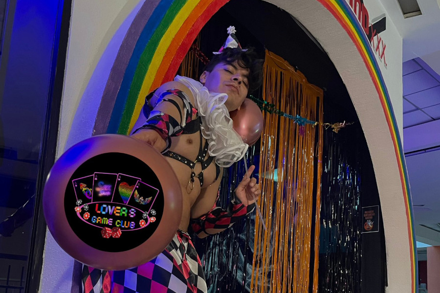 Nath BarbieBoy con un colorido traje de arlequín, de pie en la entrada de Halloween de Lovers Game Club.