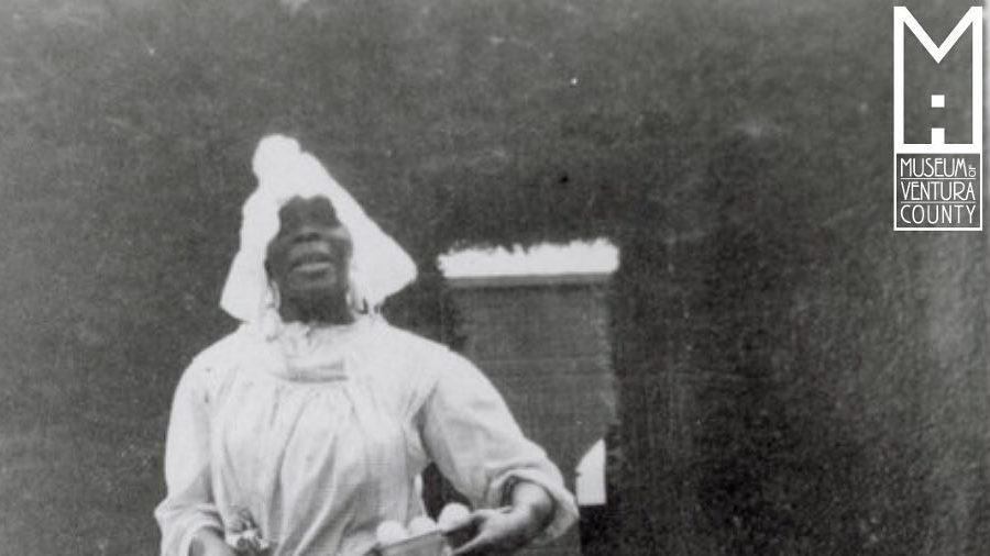 Historical black and white photo of Lucy Hicks Anderson, wearing a dress and head wrap while smiling outdoors.