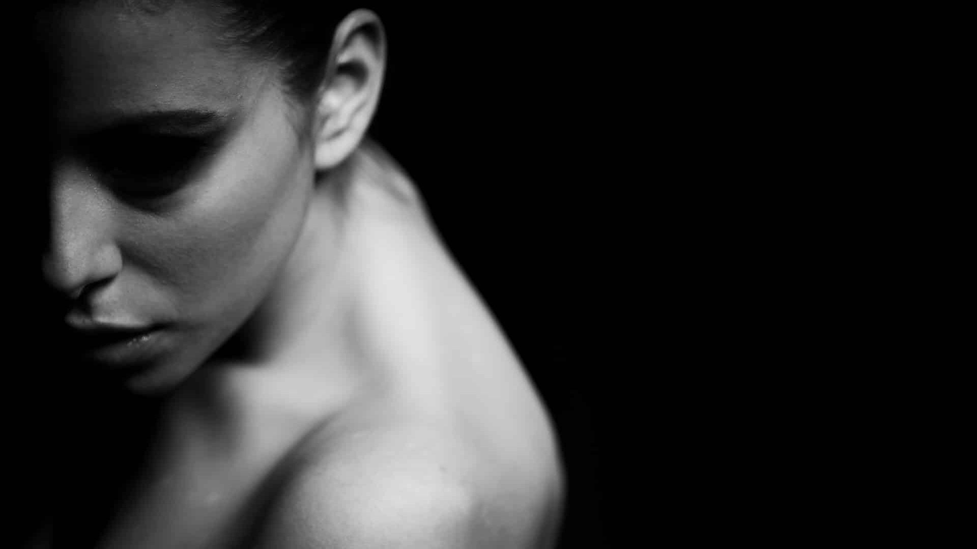 Primer plano dramático en blanco y negro del rostro y el hombro desnudo de una mujer, parcialmente en sombra.