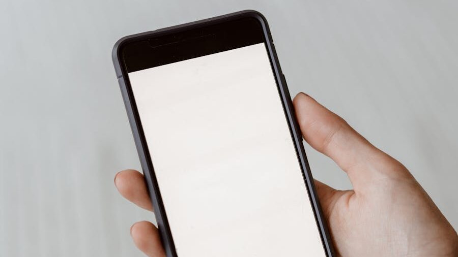 A hand holds a black smartphone with a completely blank white screen over a white textured bedspread.