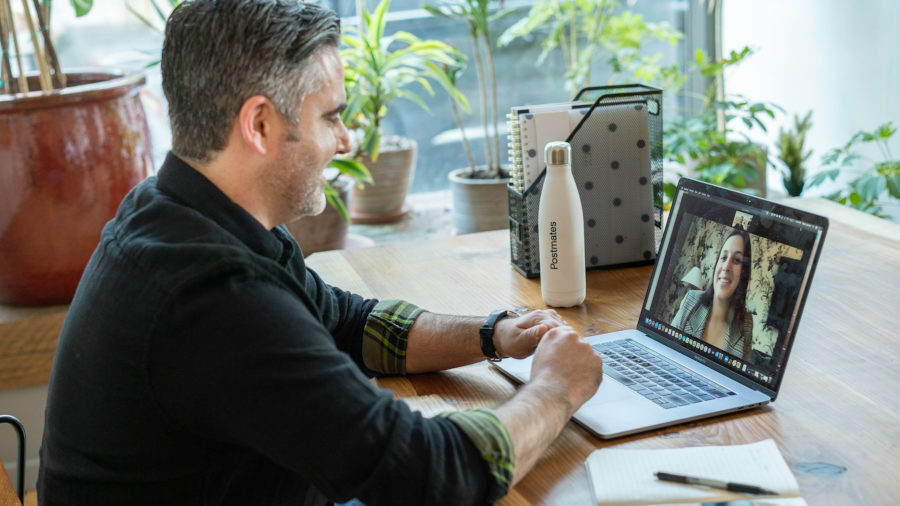 A man sits at a wooden desk smiling during a video call with a woman on his laptop screen, surrounded by notebooks and plants.