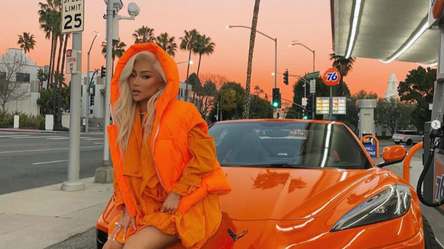 Nikita Dragun en tenue orange vif, assise sur le capot d’une Corvette assortie à Los Angeles.