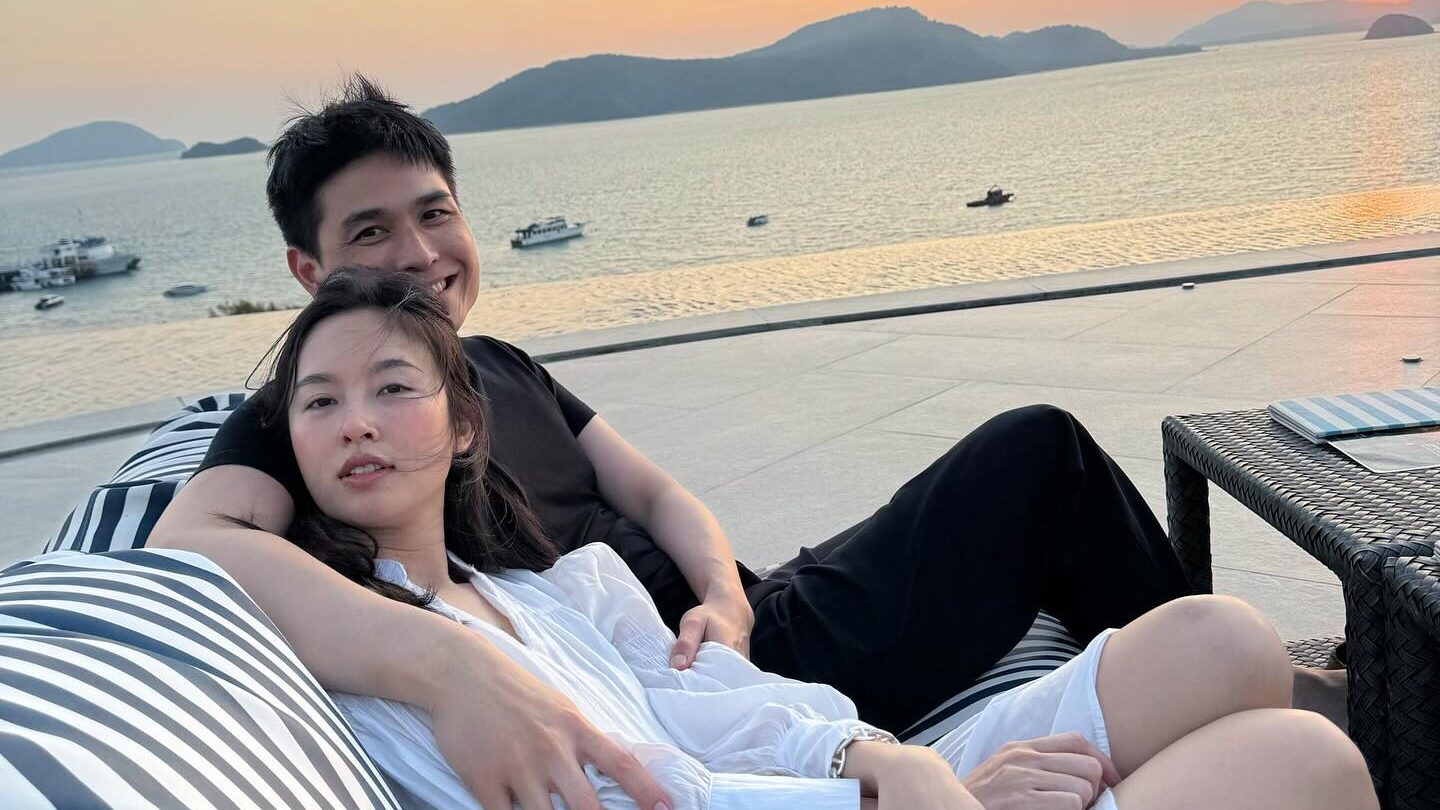 Nong Poy and her husband sit on striped cushions, hugging by the sea during a sunset.