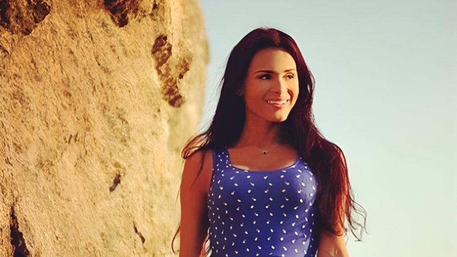 Patricia Araujo stands outdoors by a rock wall in Rio, wearing a sleeveless blue dress with white polka dots, smiling at sunset