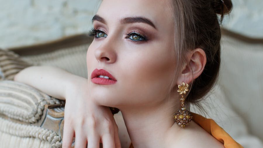 Woman with light eyes and red lipstick poses in profile, wearing gold floral earrings and soft orange dress.